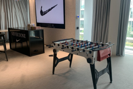 A foosball table stands before a large screen showcasing a swoosh design, while on the left, a bar counter boldly declares "NEVER DONE." The playful atmosphere hints at the lively energy of foot pool games.