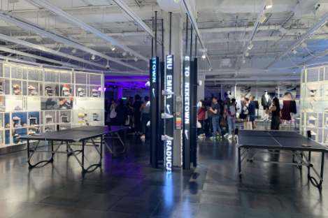 A modern indoor space with tables and illuminated signs, where a lively crowd gathers in the background near displays. Fluorescent lights hang from the ceiling, casting a glow over an intriguing foot pool area that invites playful interaction.