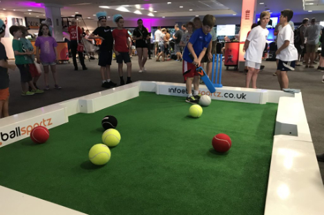 Children playing foot pool, a game combining soccer and billiards. They are kicking large tennis balls into pockets on a green rectangular platform, while bystanders look on.