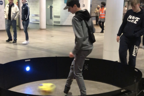 A person in a tracksuit demonstrates foot pool with a robotic device inside a circular enclosure in a public space, capturing the attention of onlookers.
