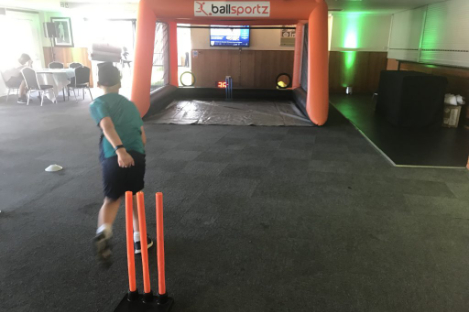 A person bowls a ball in an indoor cricket game setup with an inflatable orange frame labeled "ballsportz," reminiscent of the vibrant setting of foot pool.