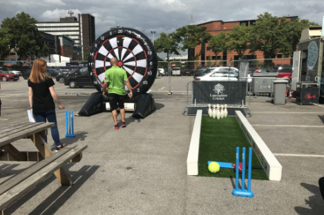 A man and woman enjoy outdoor games, including foot pool alongside a giant dartboard and miniature cricket, in a parking lot.