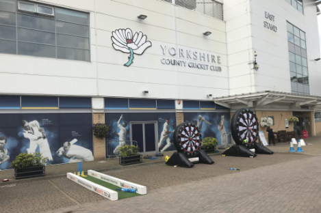 Facade of Yorkshire County Cricket Club's East Stand with inflatable dartboards, a mini-golf setup, and a lively foot pool game in front.