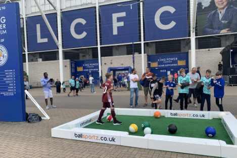 A child enthusiastically plays foot pool outside a stadium, drawing the attention of a group of onlookers. Large letters "LCFC" prominently adorn the building in the background.
