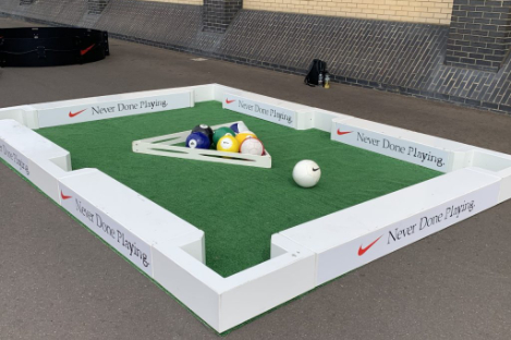 A square outdoor foot pool game with Nike branding, featuring large colored balls on a green turf surface and a white frame.