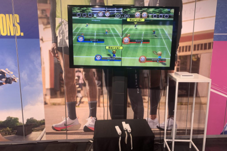 A TV screen showcases a tennis video game, with two controllers resting on a stand below, just like how foot pool pairs the strategy of pool with the intricacies of soccer.