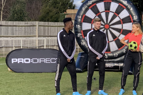 Three people in sports tracksuits stand in front of a giant inflatable dartboard on a grassy field, ready for an intense game of foot pool. They hold a soccer ball with precision, as the "PRO:DIRECT" banner flutters proudly in the background.