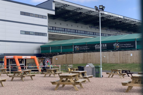 Outdoor seating area with wooden picnic tables in front of a large building and a tented fan zone. A foot pool game adds excitement alongside the inflatable game visible on the left.