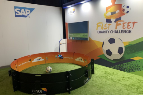 Indoor setup for a charity challenge featuring a small fenced area designed like a foot pool, complete with a soccer ball inside and vibrant event branding on the walls.
