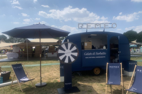 A blue gelato and sorbetto trailer with a spinning wheel and deck chairs in front is set at an outdoor event under a partly cloudy sky, right beside a foot pool for added fun.