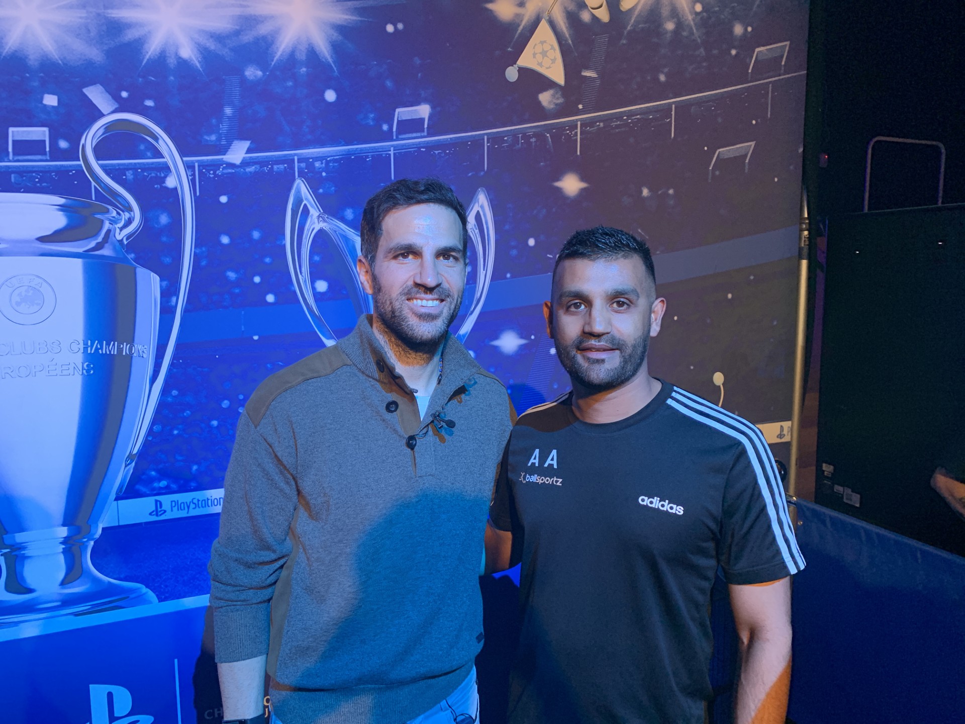 Two men stand before a UEFA Champions League trophy display. The man on the left sports a grey sweater, while the man on the right is clad in a black Adidas shirt, embodying the spirit of branded games within this prestigious football arena.