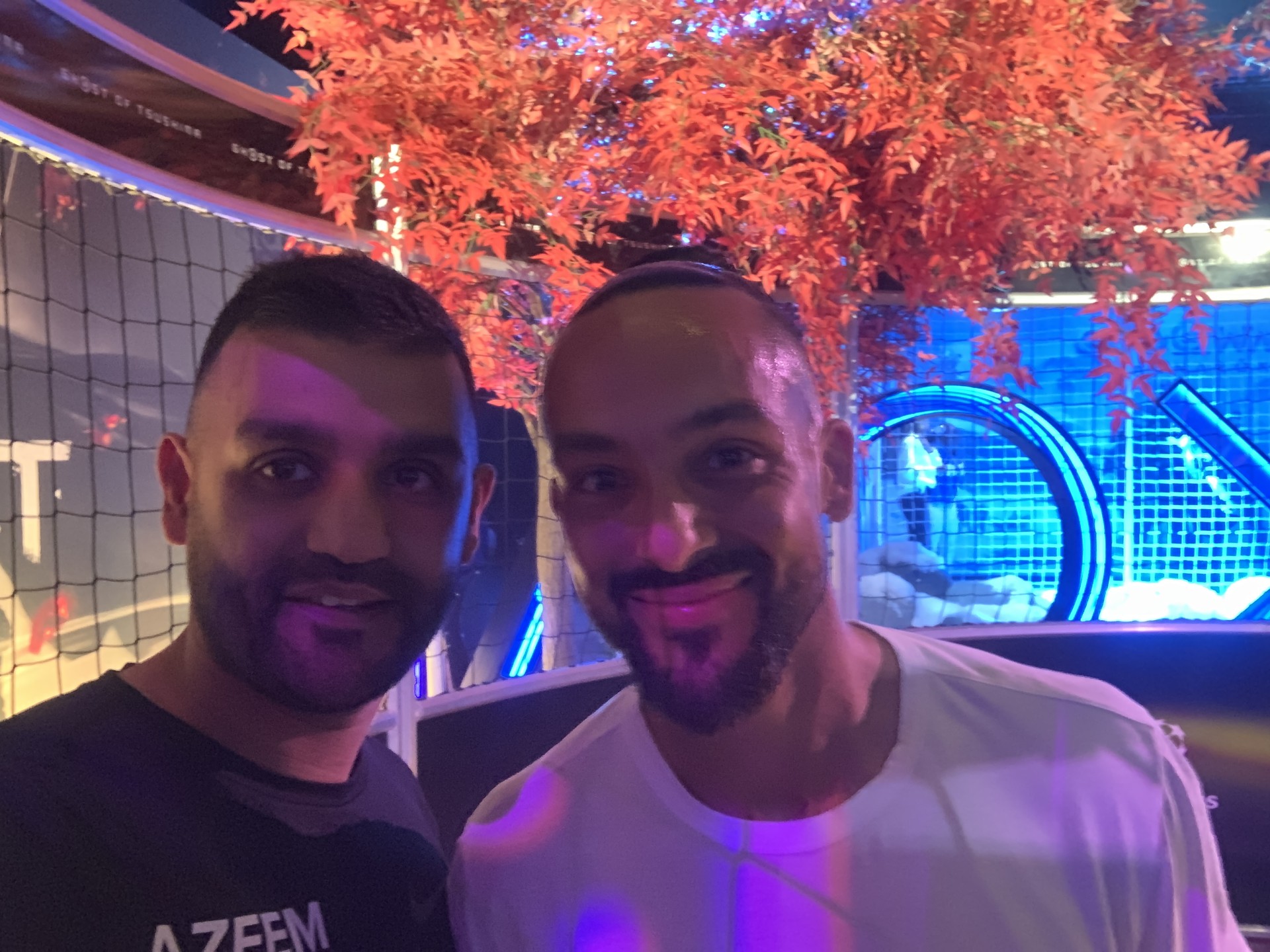 Two men capture a selfie indoors, framed by vibrant orange leaves in the background, reminiscent of the vivid graphics in branded games.