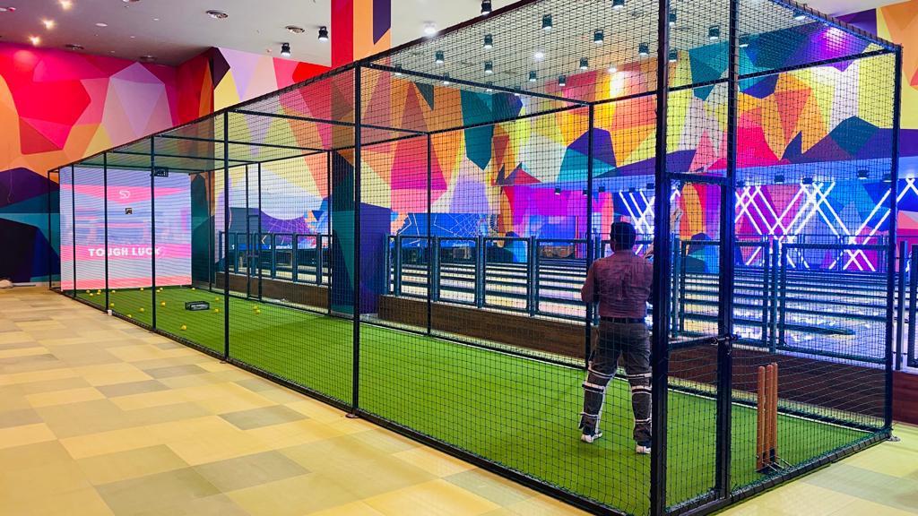 Indoor cricket practice area with netting features a mannequin in full gear at one end. Colorful patterned walls and bowling lanes create an immersive atmosphere, ideal for those seeking Cricket Simulator Hire to refine their skills.