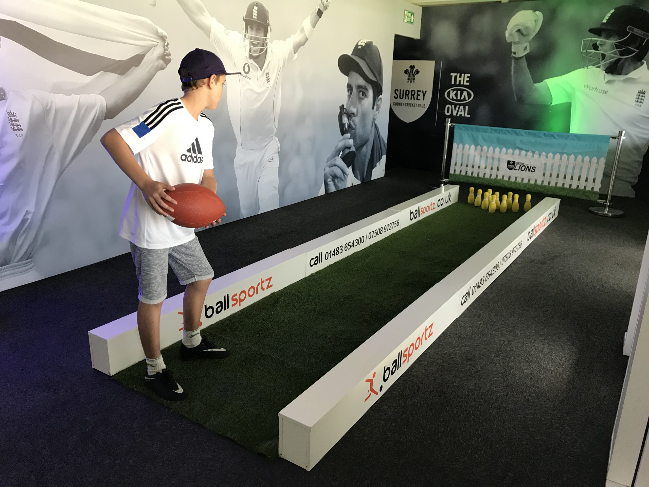 A person prepares to throw an American football at pins set up like a bowling game in a vibrant indoor sports-themed area, where cricket images adorn the walls and a foot pool game adds to the playful atmosphere.