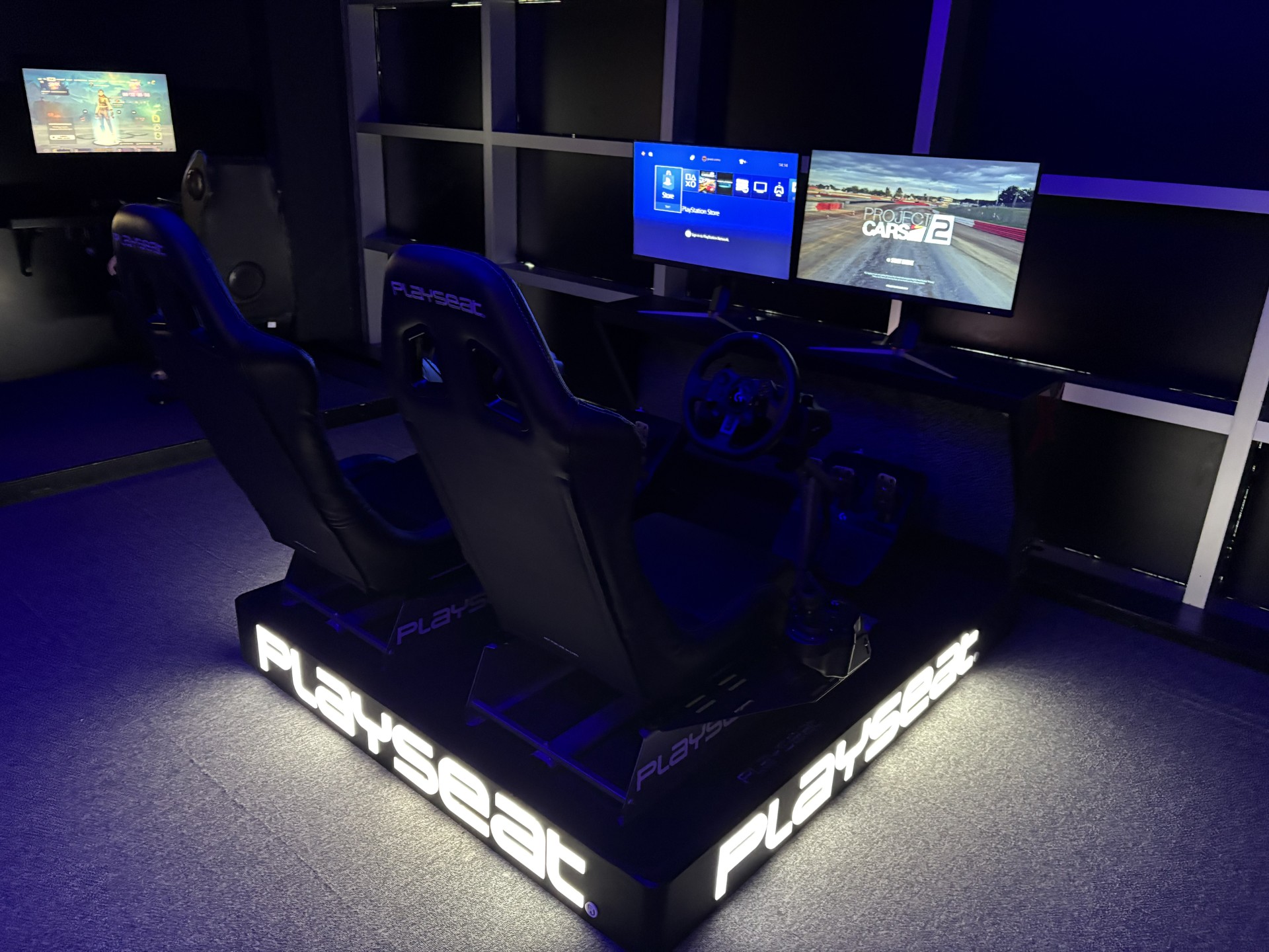 A dimly lit room features two racing simulator seats with steering wheels and pedals, facing dual monitors displaying game interfaces—a thrilling setup perfect for any event alongside a photo booth hire.