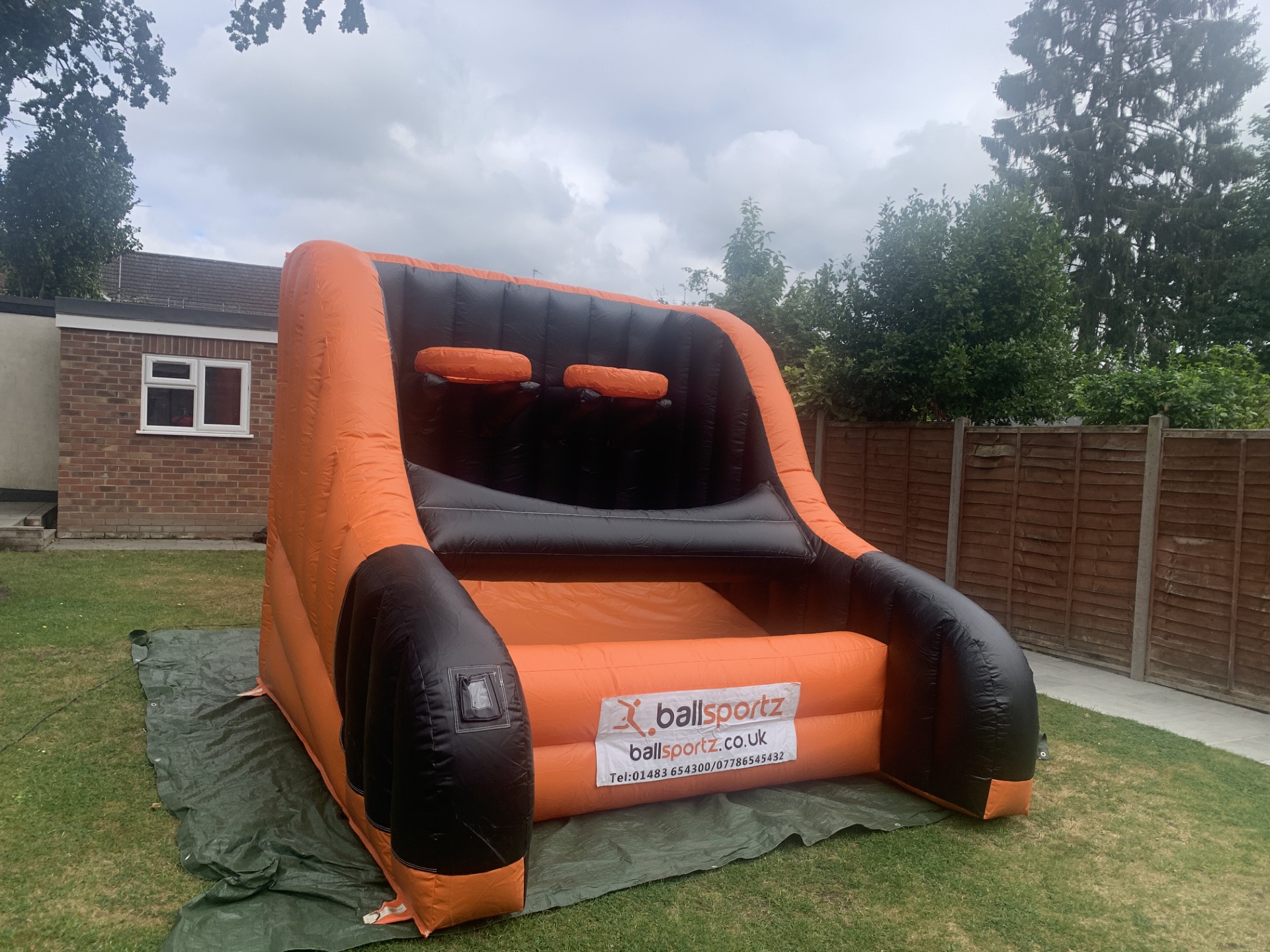 An inflatable basketball hoop game, available for hire, is set up on a lush lawn with a charming house and sturdy fence in the background.