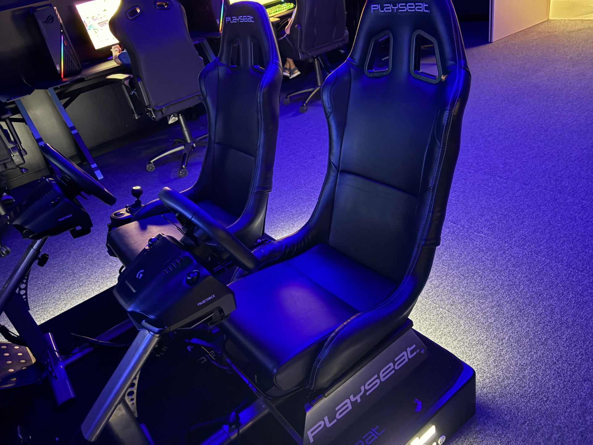 A pair of sleek black gaming seats with attached steering wheels and pedals bask in blue lighting, creating an immersive racing experience reminiscent of a high-octane arcade setting. Perfect for any event, these setups are as thrilling as a photo booth hire moment captured in time.