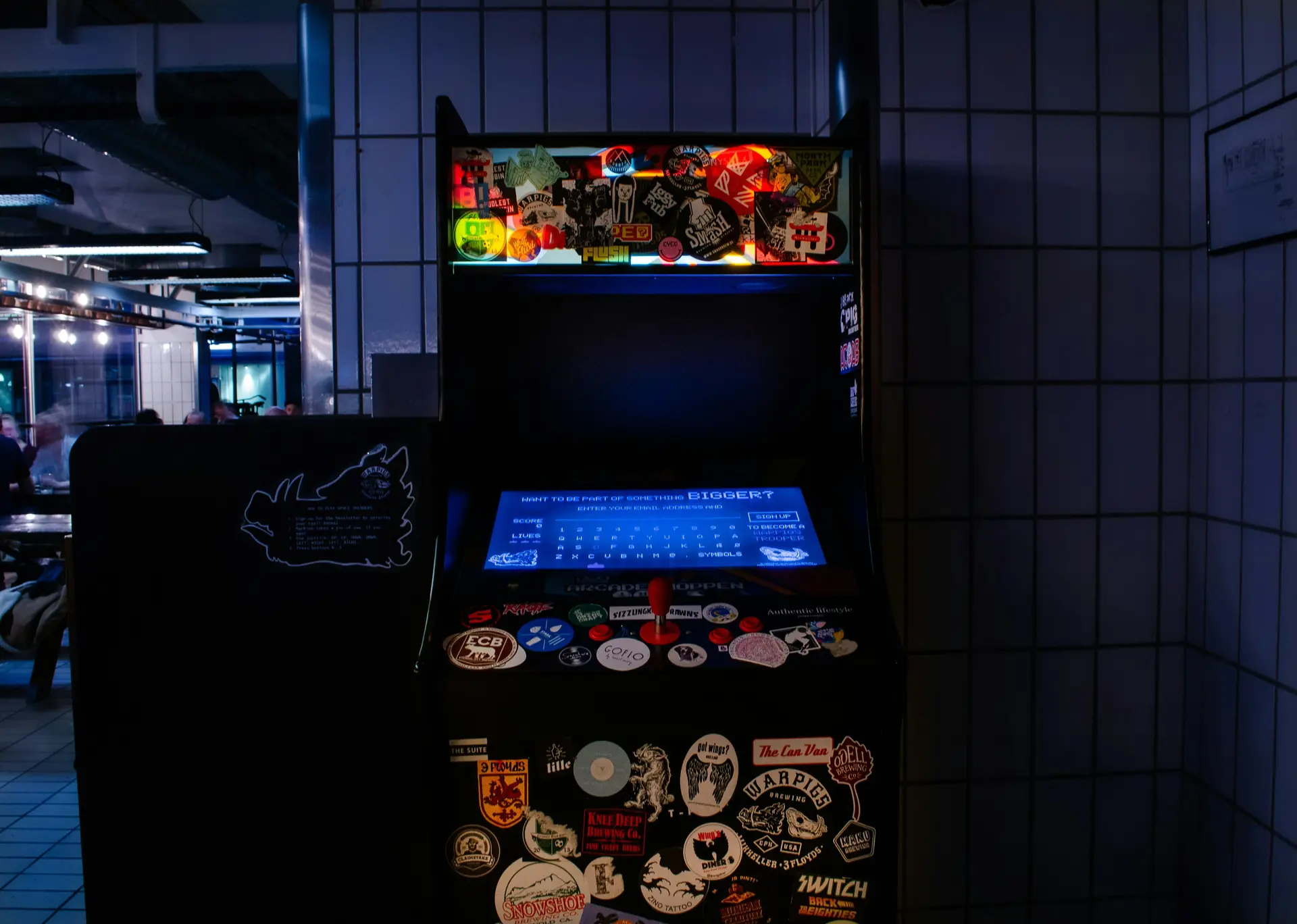 An Arcade Games Hire machine covered with stickers stands in a dimly lit tiled room.