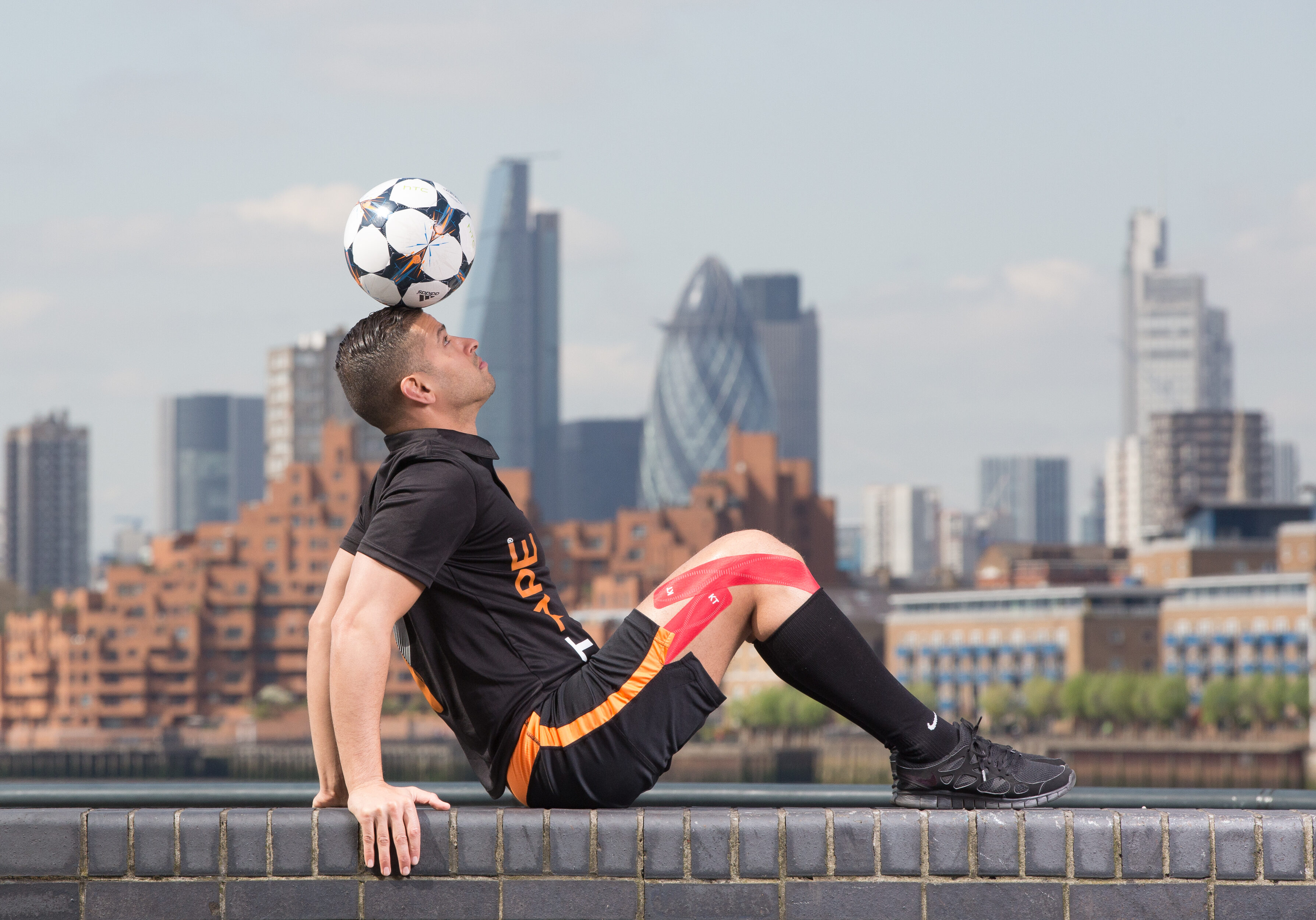 Person balances a soccer ball on their forehead while sitting on a ledge, with a cityscape in the background.