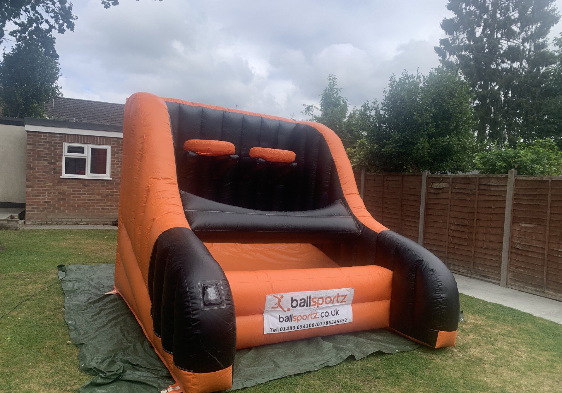 An inflatable basketball hoop game, available for hire, is set up on a lush lawn with a charming house and sturdy fence in the background.