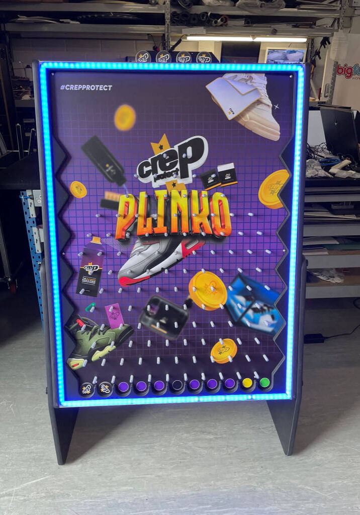 A Crep Protect Plinko board with shoes and game elements displayed, surrounded by blue LED lights, creates an electrifying ambiance perfect for your next event. Elevate the excitement with a Plink Game Hire experience that will captivate guests and add a dynamic twist to any gathering.