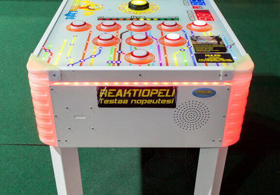 Arcade game machine with buttons and lights, labeled "REAKTIOPELI testaa nopeutesi," placed on a green carpeted floor.