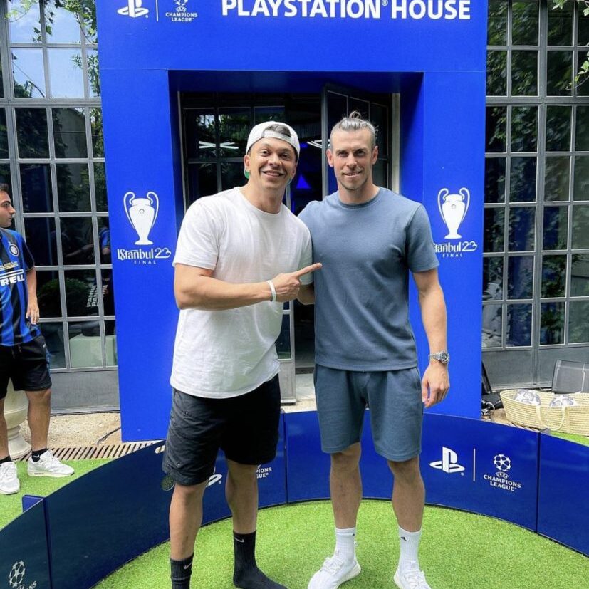Two people stand in front of a blue PlayStation House backdrop. One person is wearing a white shirt and cap, and the other is in a gray shirt and shorts.