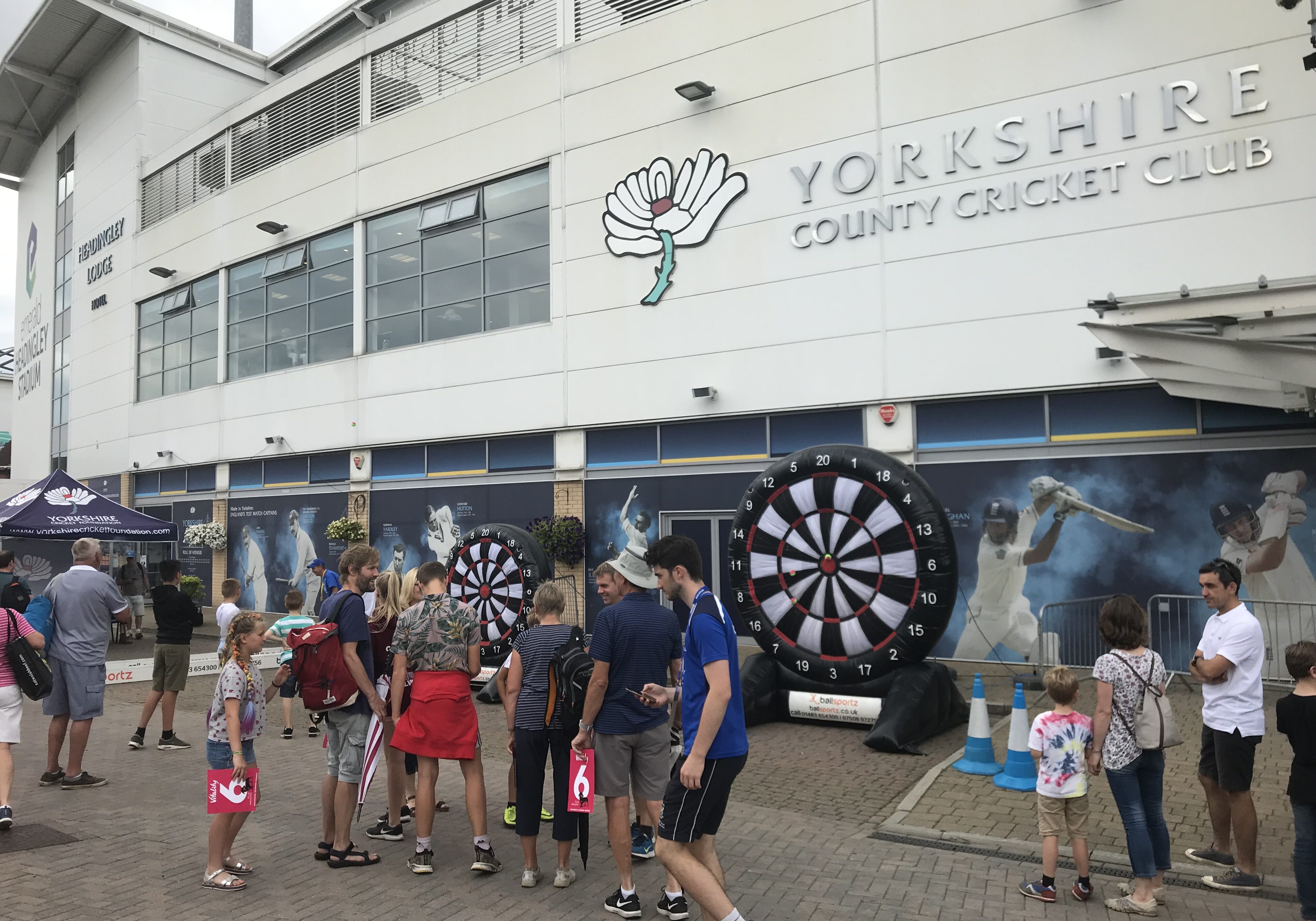 People gather outside Yorkshire County Cricket Club, with two large dartboards set up for an event.