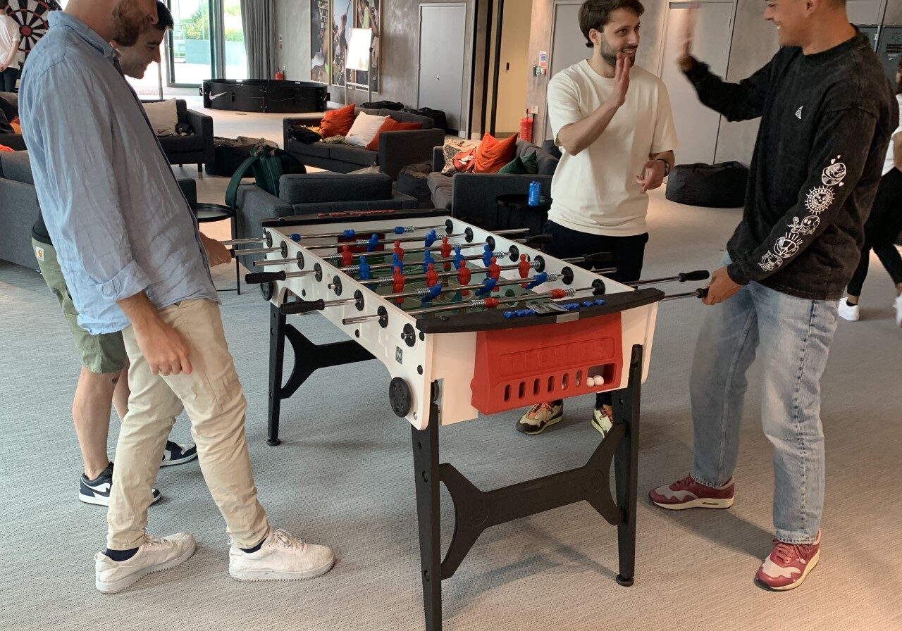 Four people playing foosball in a room with couches and a dartboard.