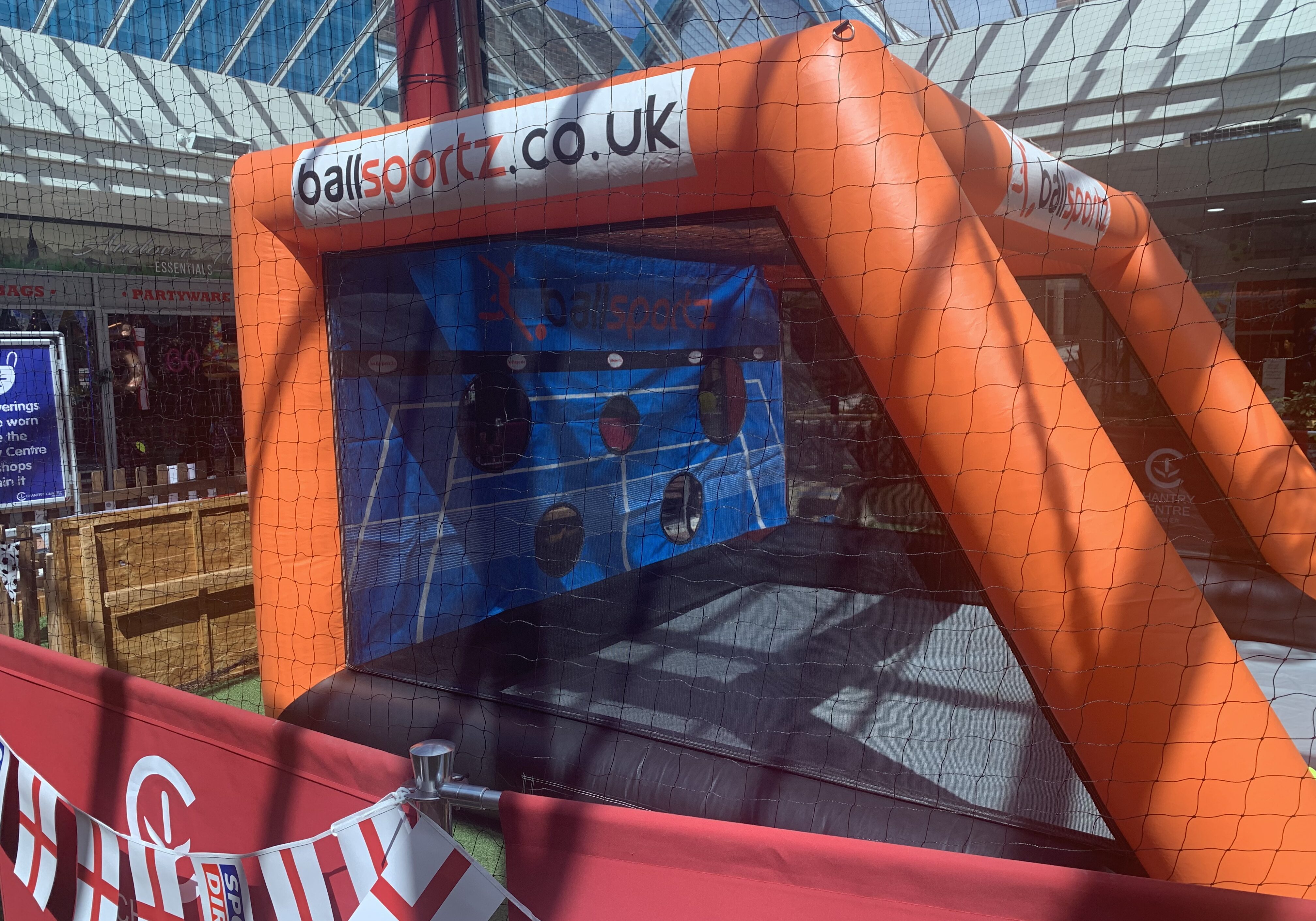 Inflatable sports game setup with BallSportz branding, featuring a netted enclosure and target holes, placed indoors with a red barrier in front.