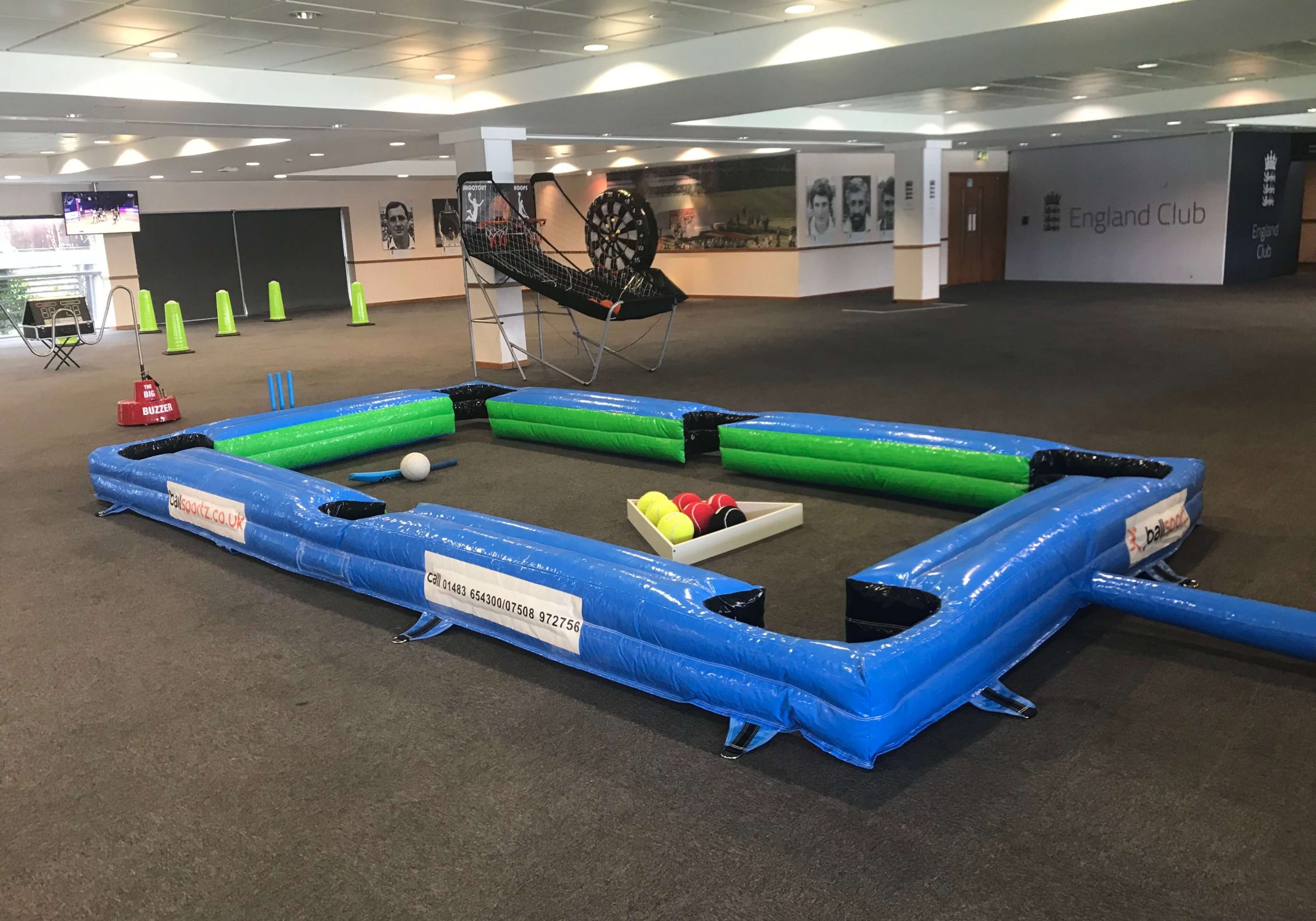 Inflatable billiards-style soccer game with a blue and green border, set up in a large indoor area with gray carpet and sports decor.