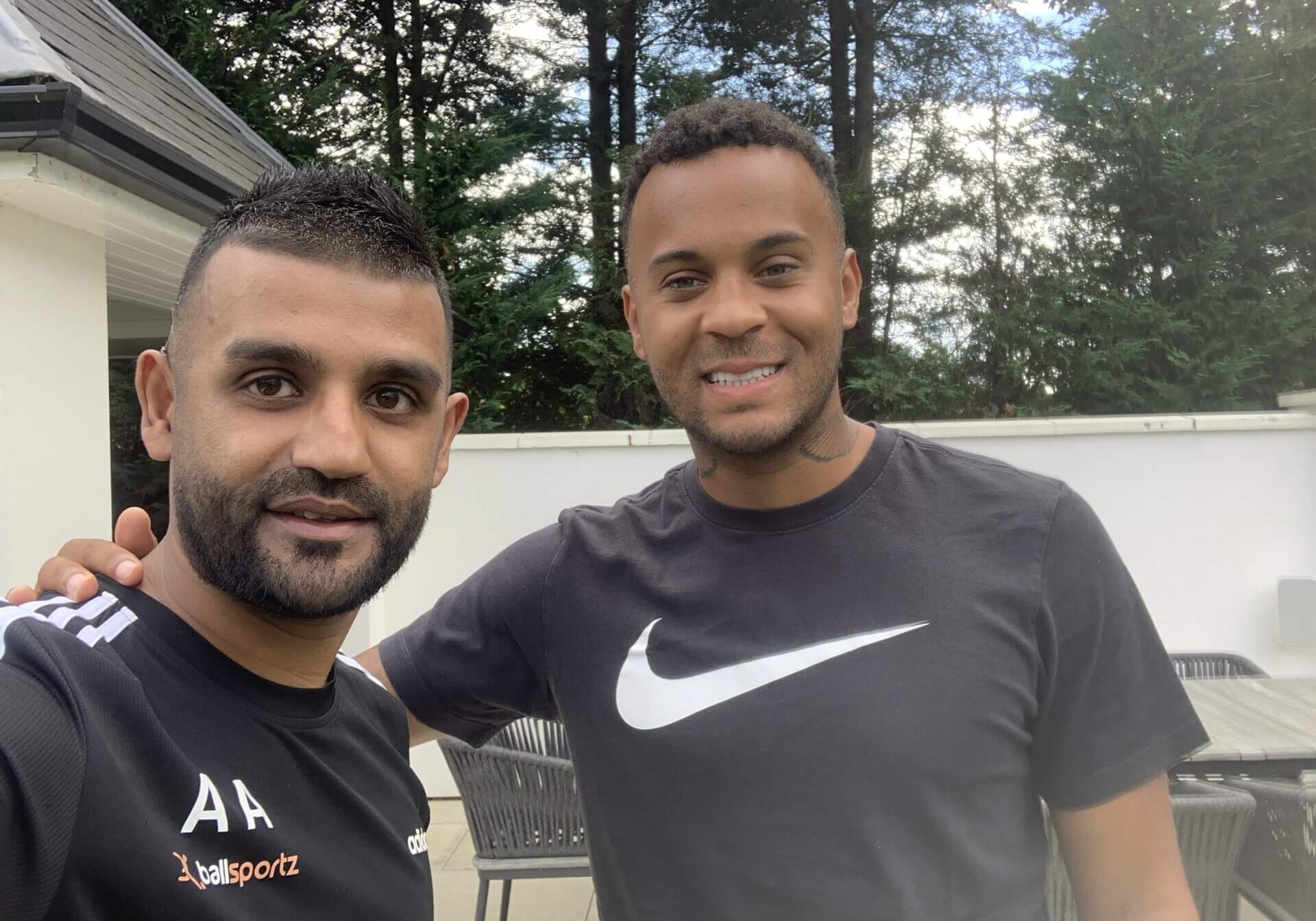 Two men standing outdoors, smiling at the camera. The man on the left wears a black shirt with "AAsports" text, while the man on the right wears a Nike T-shirt. Trees are visible in the background.